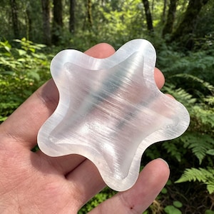 May include: A star-shaped selenite crystal carving with a smooth, translucent white surface and a fibrous, pearlescent texture. The object features five rounded points and a polished finish that reflects light across its layered, crystalline structure.