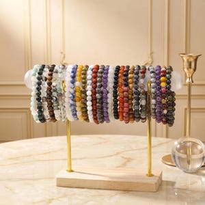 Könnte beinhalten: Eine Präsentation von Armbändern mit Perlen in verschiedenen Farben, darunter Weiß, Schwarz, Blau, Gelb, Rot und Lila. Die Armbänder sind auf einem goldfarbenen Metallständer mit Holzsockel angeordnet. Im Hintergrund ein goldener Kerzenhalter und eine marmorierte Oberfläche.