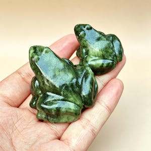 Può includere: Due piccole statuette di rana in giada verde. Le rane sono scolpite con dettagli e sono fianco a fianco. La giada ha un aspetto screziato con variazioni di colore verde. Le statuette sono tenute in una mano.