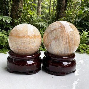 May include: Two polished, spherical quartz stones with translucent white and golden-brown inclusions. Each smooth, rounded stone rests securely on a circular, dark brown, high-gloss wooden display stand with a carved, tiered base design.