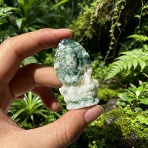 Può includere: Una piccola statuetta in pietra intagliata a mano che raffigura una divinità seduta con testa di elefante, caratterizzata da un motivo maculato di verde bosco intenso e inclusioni minerali bianco traslucido sulla superficie liscia.