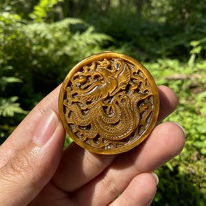 May include: A circular tiger's eye gemstone pendant featuring an intricate, openwork carving of a phoenix with detailed wings and a long, flowing tail. The stone displays a characteristic golden-brown chatoyant lustre with natural banding.