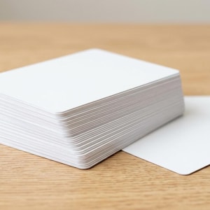 Puede incluir: Una pila gruesa de tarjetas rectangulares blancas con esquinas redondeadas, acompañada de dos tarjetas blancas sueltas colocadas planas junto al montón principal. Las tarjetas tienen un acabado mate liso y son de tamaño uniforme.