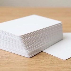 May include: A thick stack of rectangular white cards with rounded corners, arranged in a neat pile. Beside the main stack, two additional individual white cards are lying flat, showcasing the same plain, blank surface and consistent card stock material.