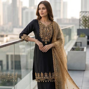 Puede incluir: Salwar kameez negro bordado con detalles dorados. El conjunto incluye un dupatta a juego y pantalones palazzo. El escote, las mangas y el dobladillo están adornados con intrincados bordados dorados. La palabra "CELESTRA" es visible en la parte inferior.