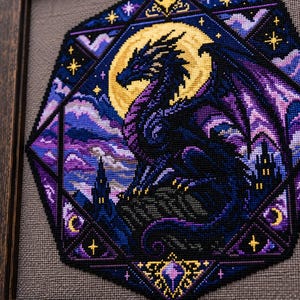 May include: A framed cross-stitch embroidery featuring a dark blue and purple dragon perched on a rocky outcrop against a large yellow moon. The scene is enclosed in a hexagonal border with starry accents, purple clouds, and silhouettes of castle towers.