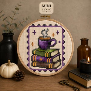 May include: A circular wooden embroidery hoop containing a cross-stitch design of a purple mug with steam resting on a stack of three books. The design features a purple and black scalloped border with small orange and purple stars. Text reads MINI 2.2" x 2.6".