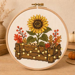 Puede incluir: Un bastidor de bordado circular de madera con un patrón de punto de cruz de un gran girasol amarillo y hojas de otoño en una caja de madera marrón. La caja está decorada con una cadena de pequeñas luces blancas sobre tela blanca.