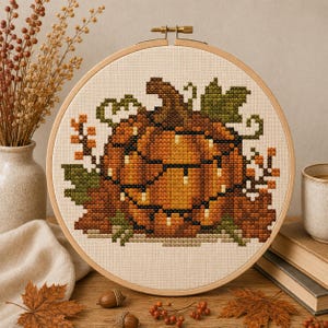 Puede incluir: Un proyecto de bordado en punto de cruz en un bastidor de madera redondo que muestra una gran calabaza naranja envuelta en una cadena de pequeñas luces blancas, rodeada de hojas verdes, follaje marrón y pequeñas bayas naranjas sobre tela crema.