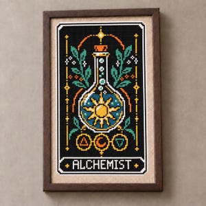 Könnte beinhalten: Ein gerahmtes Kreuzstich-Stickbild auf beigem Stoffhintergrund mit einem schwarzen rechteckigen Feld, das eine zentrale Glasflasche mit einer gelben Sonne, grünen Blättern und drei kreisförmigen Symbolen zeigt, sowie dem Wort ALCHEMIST unten.