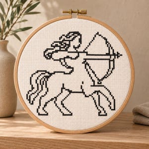 Könnte beinhalten: Ein runder hölzerner Stickrahmen mit weißem Stoff, der ein schwarzes Kreuzstichmotiv eines Zentaur-Bogenschützen zeigt. Der Zentaur ist im Profil dargestellt, wie er mit Pfeil und Bogen zielt, und hat eine fließende Mähne sowie einen Schweif.