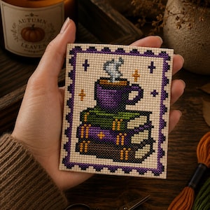 May include: A rectangular cross-stitch embroidery piece featuring a purple mug with steaming liquid resting on a stack of three books. The design is bordered by a purple and black scalloped frame on a cream-colored fabric background with small star accents.