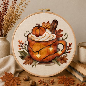 Puede incluir: Un bastidor de bordado circular de madera que contiene un patrón de punto de cruz de una taza naranja llena de nata montada, una calabaza y ramas de canela. El diseño presenta hojas de otoño, enredaderas y bayas en tonos naranja, marrón y verde.