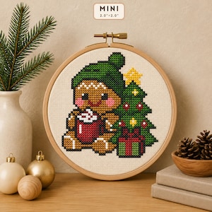 Puede incluir: Un patrón de punto de cruz en un bastidor de madera redondo que muestra un hombre de jengibre con gorro verde, sosteniendo una taza roja con espuma blanca, junto a un árbol de Navidad decorado y un regalo, con la etiqueta MINI 5,1cm x 5,1cm.
