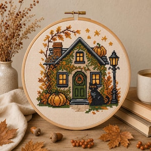 May include: A circular wooden embroidery hoop containing a cross-stitch pattern of a house covered in autumn foliage. The design features a green door, glowing yellow windows, orange pumpkins, a black cat, and a black street lamp on cream fabric.