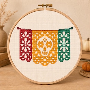 May include: A circular wooden embroidery hoop containing a cross-stitch design on white fabric. The pattern features three vertical banners in red, yellow, and green, with a white skull centered on the yellow banner and floral motifs.