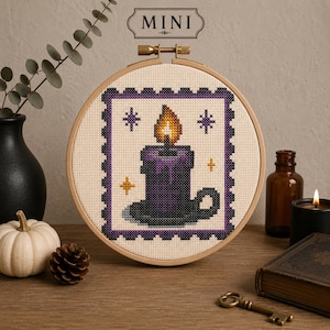 May include: A circular wooden embroidery hoop containing a cross-stitch design of a purple candle in a holder with a yellow flame. The design is framed by a purple and black scalloped border on cream fabric. The text MINI is printed above the hoop.