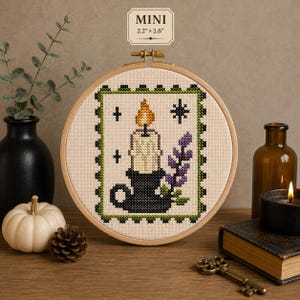 Puede incluir: Un bordado de punto de cruz en un bastidor de madera redondo con una vela en un soporte negro y una rama de flores moradas. El diseño incluye un borde festoneado verde y estrellas. Una etiqueta arriba indica MINI 5,6 cm x 6,6 cm.