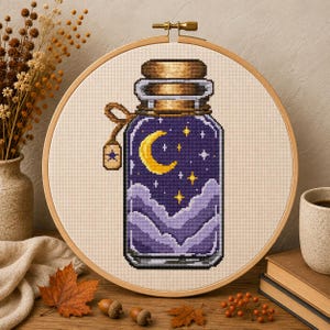 Puede incluir: Un bordado de punto de cruz en un bastidor de madera redondo que muestra una botella de vidrio llena con una escena de cielo nocturno. El diseño incluye una luna creciente amarilla, estrellas blancas y amarillas, y nubes moradas. Una pequeña etiqueta con una estrella cuelga del cuello.