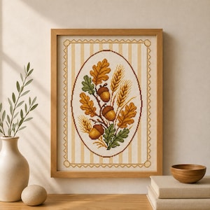 May include: A framed cross-stitch embroidery featuring an oval arrangement of brown acorns, green oak leaves, golden-brown oak leaves, and stalks of wheat. The design is set against a cream and beige vertical striped background with a scalloped border.