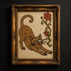 May include: A cross-stitch embroidery featuring a brown cat stretching its body, positioned next to a green vine with two orange flowers. The artwork is encased in an ornate, antique-style gold frame with intricate carved detailing on the edges.
