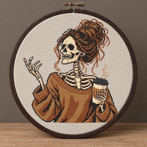 Puede incluir: Un bastidor de bordado circular con un diseño de punto de cruz de un esqueleto con cabello castaño en un moño, vistiendo un suéter marrón. El esqueleto sostiene una taza de café humeante y gesticula con la otra mano sobre tela clara.