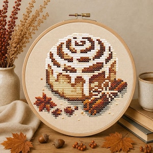 May include: A circular wooden embroidery hoop containing a cross-stitch pattern of a cinnamon roll with white icing, accompanied by a cinnamon stick and star anise, all set against a light beige fabric background with a visible grid texture.