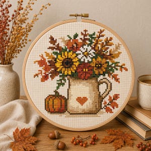 Puede incluir: Un bastidor de bordado circular de madera que contiene un patrón de punto de cruz de una jarra beige con un corazón, llena de girasoles, margaritas blancas y hojas de otoño, acompañada de una pequeña calabaza naranja sobre tela color crema.