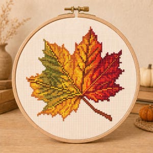 Puede incluir: Un bordado en punto de cruz de una hoja de arce centrada en un bastidor de madera redondo. La hoja presenta un degradado de colores que va del verde a la izquierda al amarillo en el centro y al rojo oscuro a la derecha sobre tela blanca.