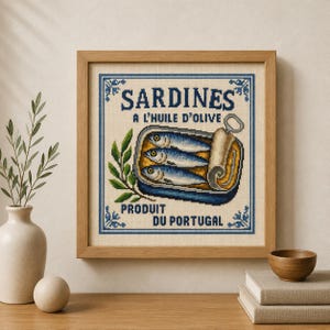 Może przedstawiać: Kwadratowy haft krzyżykowy w drewnianej ramie przedstawiający otwartą puszkę z trzema niebiesko-srebrnymi sardynkami. Tekst głosi SARDINES A L'HUILE D'OLIVE oraz PRODUIT DU PORTUGAL. Po lewej stronie puszki znajduje się zielona gałązka oliwna.