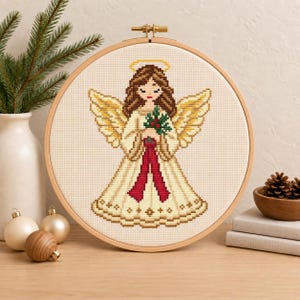 May include: A cross-stitch embroidery of an angel with brown hair, a yellow halo, and large golden wings, wearing a cream-colored gown with a red ribbon. The angel holds a sprig of green holly with red berries. The design is framed in a wooden hoop.