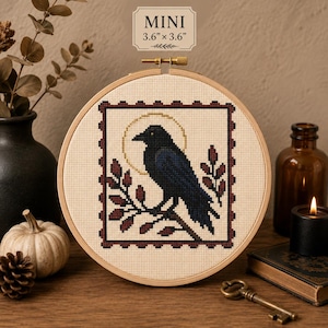May include: A circular wooden embroidery hoop containing a cross-stitch design of a black crow perched on a branch, framed by a dark red border. Above the crow is a gold halo. The fabric is cream-colored. Text above reads MINI 3.6" x 3.6".