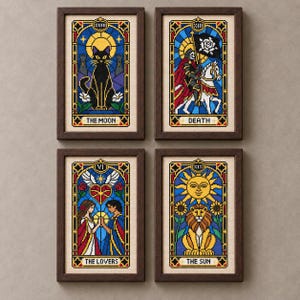 May include: Four framed cross-stitch artworks depicting tarot cards. The top left shows a black cat labelled THE MOON. The top right shows a figure on a horse labelled DEATH. The bottom left shows two figures labelled THE LOVERS. The bottom right shows a lion labelled THE SUN.