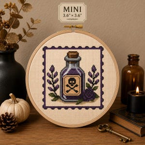 May include: A round wooden embroidery hoop containing a cross-stitch design of a purple potion bottle with a skull and crossbones label, flanked by lavender sprigs, framed by a purple border. Text above reads MINI 3.6" x 3.6".