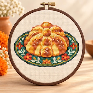 May include: A cross-stitch embroidery in a circular wooden hoop featuring a golden-brown pan dulce bread centered on a teal oval plate decorated with orange and red floral patterns against a cream-colored fabric background.