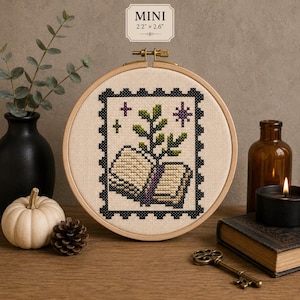 Puede incluir: Un bordado de punto de cruz en un bastidor de madera redondo que muestra un libro con una planta verde creciendo de sus páginas, enmarcado por un borde de sello postal negro con estrellas moradas. El texto MINI 5,6 cm x 6,6 cm aparece arriba.