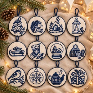 May include: Twelve circular cross-stitch ornaments featuring blue thread designs on white fabric. Patterns include an owl, hedgehog, house, lantern, sled, stocking, snow globe, winter hat, bird, snowflake, teapot, and a gift box, each with a dark blue loop.
