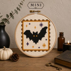 Può includere: Un telaio da ricamo circolare in legno contenente un motivo a punto croce di un pipistrello nero con occhi gialli, circondato da piccole stelle viola e arancioni, incorniciato da un bordo a francobollo. La parola MINI è stampata sopra il telaio.