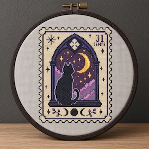 Puede incluir: Un bordado de punto de cruz en un bastidor de madera oscura con diseño de sello postal. Muestra un gato negro sentado en una ventana gótica mirando una luna creciente amarilla y estrellas en un cielo morado, con el texto 31 CEMTS.