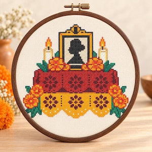 May include: A cross-stitch embroidery in a round wooden hoop featuring a central framed silhouette portrait, flanked by two lit candles, resting on a red and yellow altar decorated with orange marigold flowers and green leaves.