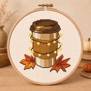 May include: A cross-stitch embroidery in a round wooden hoop featuring a brown coffee cup wrapped in a string of glowing yellow fairy lights, positioned between two orange and brown autumn maple leaves on a white fabric background.
