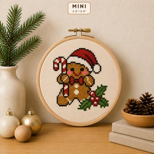 Puede incluir: Un bastidor de bordado circular de madera con un diseño de punto de cruz de un hombre de jengibre con un gorro de Papá Noel rojo y blanco, sosteniendo un bastón de caramelo junto a hojas de acebo. El texto MINI 5,1 cm x 5,1 cm es visible.