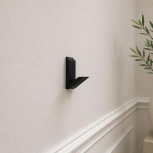 Può includere: Un gancio da parete nero opaco con un design geometrico e angolare. Il gancio presenta una piastra di base rettangolare verticale e un braccio sporgente che si estende verso l'esterno con un'angolazione verso l'alto, offrendo un punto di sospensione.