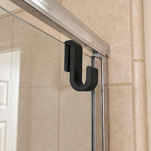May include: A matte black over-the-door hook designed for a glass shower enclosure. The hook features a U-shaped bracket that fits securely over the top frame of the shower door, providing a sturdy hanging point for towels or bathroom accessories.
