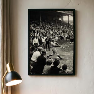 Può includere: Una fotografia incorniciata in bianco e nero che ritrae un giocatore di baseball in divisa a righe mentre colpisce una palla con una mazza durante una partita. La scena include una grande folla di spettatori sugli spalti e persone a bordo campo.