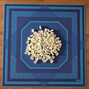 May include: A blue Mahjong game mat with a geometric design and a pile of ivory-colored tiles. The mat has a dark blue border with text and a lighter blue central area. The tiles are scattered in the center.