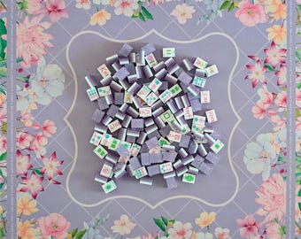 May include: A collection of purple and white Mahjong tiles scattered on a floral patterned game board. The board features a lavender background with a white diamond pattern and a border of pink and white flowers.