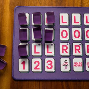 Könnte beinhalten: Ein lila Spielbrett mit Buchstaben- und Zahlenkacheln und durchscheinenden lila Spielsteinen. Das Brett trägt die Worte "BOLDE MOVE" und eine Vogelgrafik. Die Kacheln haben die Buchstaben L, O und R sowie die Zahlen 1, 2 und 3.