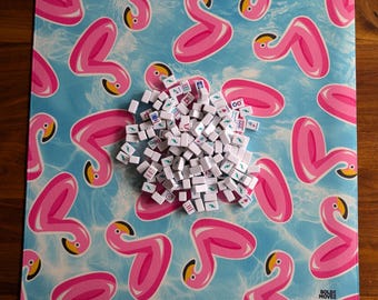 May include: A square game mat with a blue background featuring pink flamingo pool floats. A pile of white game tiles with black and green symbols sits on the mat. The mat has the text "BOLDE MOVE" in the bottom right corner.
