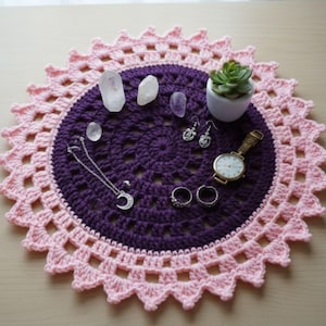 May include: A handmade crochet doily in purple and pink, with a scalloped edge. The doily holds crystals, a small succulent in a white pot, jewelry, and a gold watch.